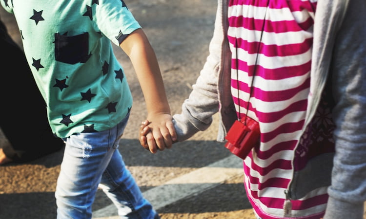 Two young students hold hands while crossing the road