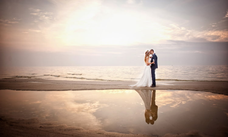 A bride and groom embrace on an empty beach, the water reflecting the sky above like a mirror